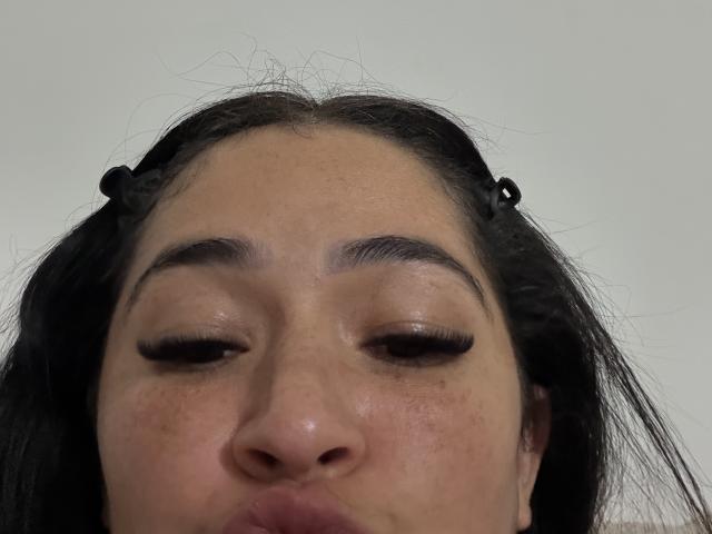 NicoColeman's face, brown eyes closed, black hair in pigtails with small black hair ties, light brown skin, subtle freckles, slightly puffy lips, neutral expression, slightly messy hair.