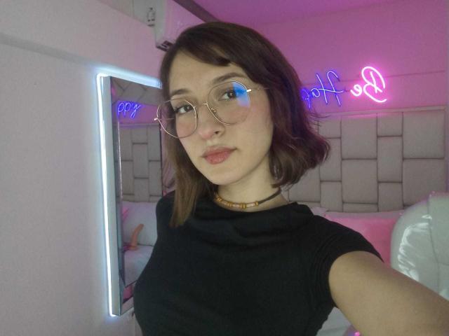CattalinaBotero has short brown hair, wears glasses, and a black shirt, with a choker, showing a subtle smile in a pink-lit room with neon "off" sign.
