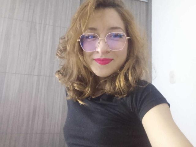 CattalinaBotero with wavy brown hair, pink-tinted sunglasses, red lipstick, and a black shirt, smiling at the camera. Boobs are not visible. Ass and pussy are not visible.