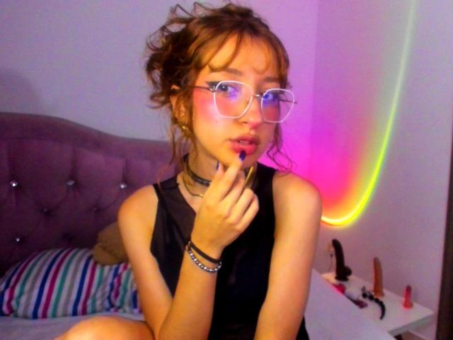 CattalinaBotero with messy brown hair, glasses, and black sleeveless top, biting finger with blue nail polish, wearing a black choker, and bracelet, sitting on a bed with sex toys.