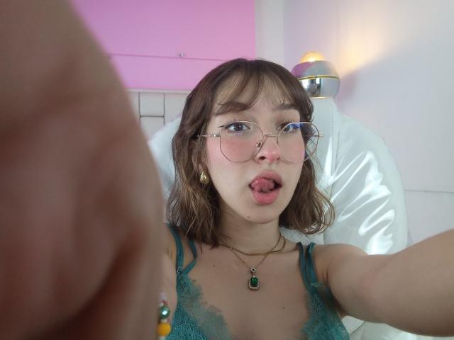 CattalinaBotero with short brown hair, glasses, green lace top, and tongue out, holding phone, necklace with green pendant, white chair, pink and white wall, small lamp in background.