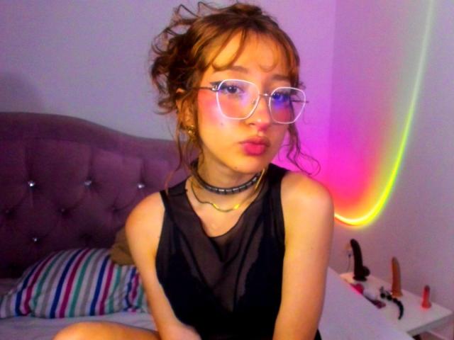 CattalinaBotero with messy brown hair, glasses, pink lips, black sheer top, choker, and striped pillow, sitting on bed with dildo and butt plug on white nightstand.