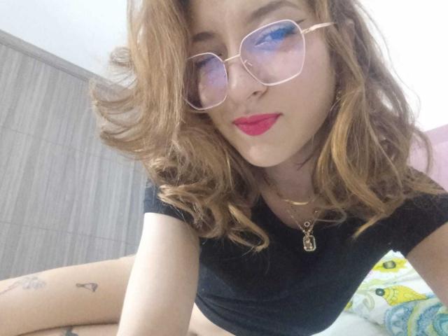 CattalinaBotero has wavy brown hair, blue-tinted glasses, red lipstick, black crop top, and a gold necklace. Her small tits and tattoo are visible. She's lying on a bed.