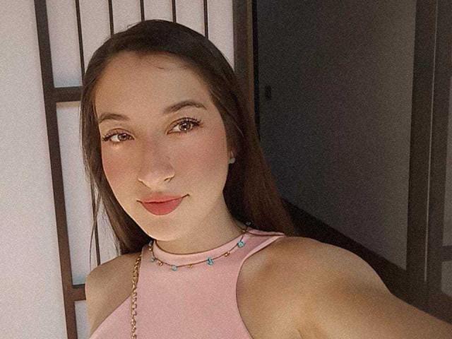 CattalinaBotero has light skin, brown eyes, and long brown hair. She's wearing a pink sleeveless top with a gold chain, and has red lipstick, smiling at the camera.