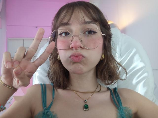 CattalinaBotero with puffy pink lips, glasses, green lace bra, and gold necklace, making a peace sign, brown wavy hair, gold earrings, and green gem necklace, looking cute.