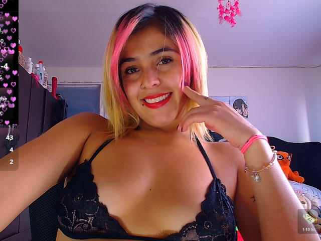 LaSexyColombiana with pink and blonde hair, smiling, wearing a black lace bra, showing her tits, pink nose ring, red lipstick, and pink bracelet, in a brightly lit room.