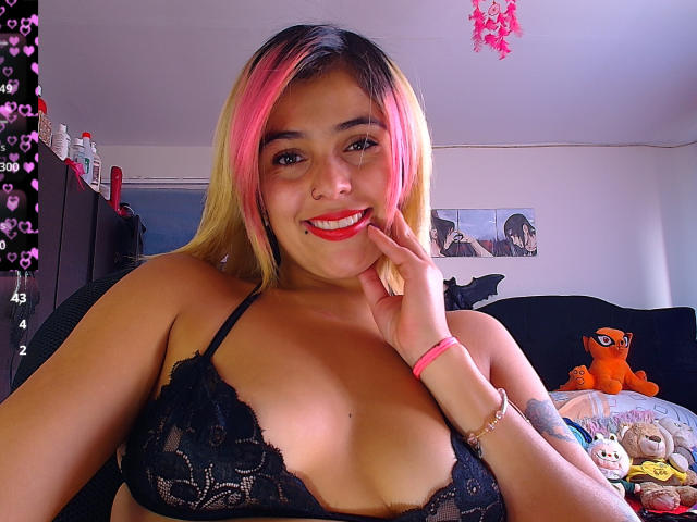 LaSexyColombiana with pink and blonde hair, smiling, wearing black lace bra, hand on cheek, red lipstick, visible tattoo on left arm, pink bracelet, and stuffed orange Pokémon on bed.