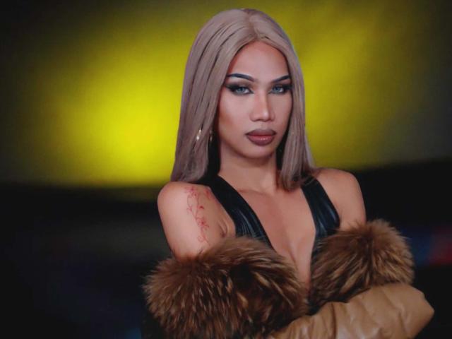 DemiAnderson with blonde hair, black leather top, fur trim, and tattoo on right shoulder, looking serious with crossed arms, yellow background. Boobs visible under leather top.