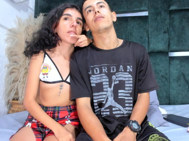 ValentinaMike with curly black hair and small tits, wearing a yellow bikini top and red plaid shorts, sucking her finger. Next to her, a bald man in a black shirt.