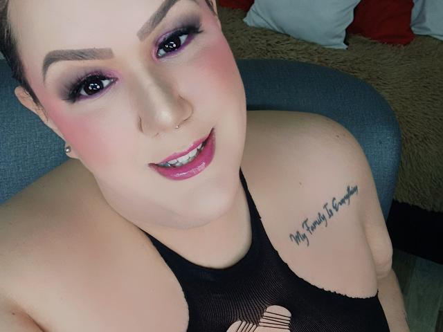 GiiannaMorrys with short brown hair, purple eye makeup, pink blush, shiny pink lipstick, black mesh top, tattoo on right shoulder, smiling, sitting on gray chair.