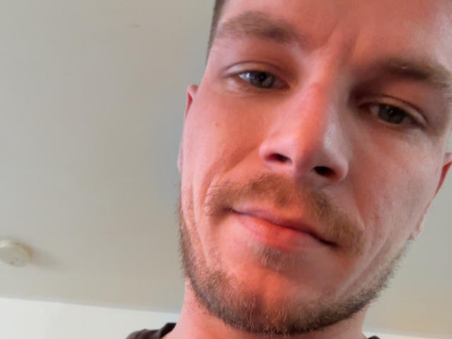 Seryll69's face, short brown hair, light beard, slight smile, blue eyes, looking at camera, slightly red skin, overhead light in background.