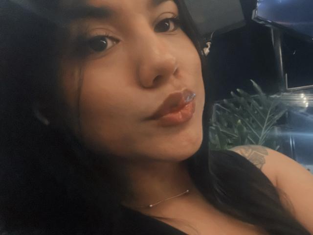 SteffanyTaylor with dark hair, brown eyes, and glossy orange lipstick, showing a tattoo on her right shoulder, wearing a black top, and a thin necklace.