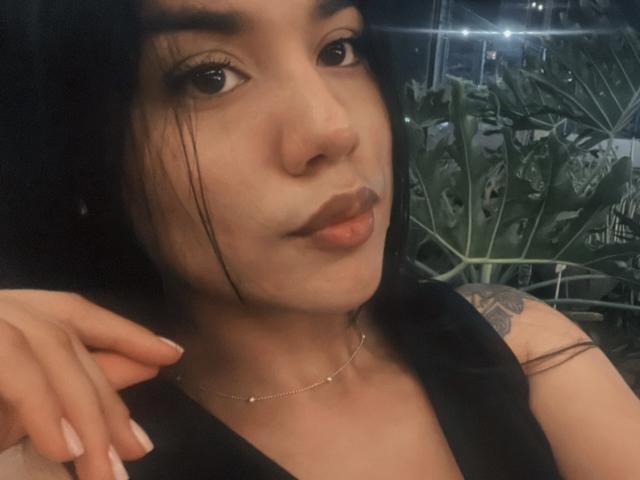 Close-up of SteffanyTaylor with dark, straight hair, slight side-swept, looking at the camera with a neutral expression. She has light brown skin, full lips, and brown eyes with subtle makeup. Her right hand is near her face, showing manicured nails with white polish. She wears a thin, silver chain necklace. A small tattoo on her upper left arm is visible. The image has a soft, slightly blurry texture. Light from the top right creates a small glare.