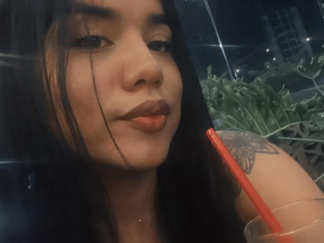 Close-up of SteffanyTaylor with long black hair, brown eyes, and dark lipstick. Her left eyebrow is slightly raised, and she's looking at the camera with a slight smile. A red straw is in her right hand, which is holding a clear plastic cup. Her left shoulder has a detailed tattoo of a flower. The image has a slight blur and a soft, dark color palette. Her skin is light brown, and she has a small nose piercing. The background is out of focus.