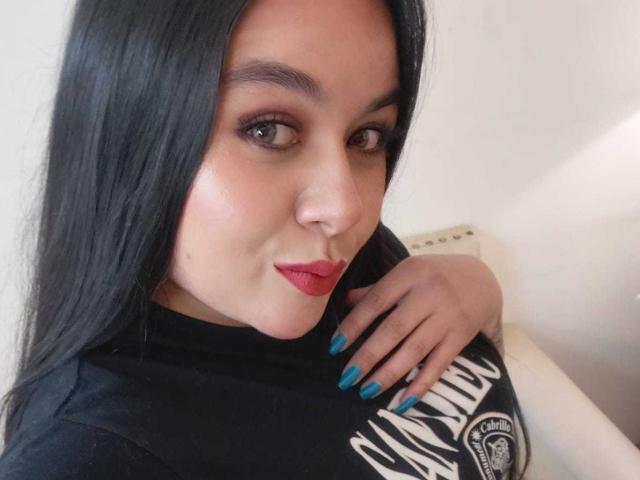 HannahMolly has black hair, red lipstick, blue nail polish, and is wearing a black shirt with white text. Her hand rests on her chest, showing her tits slightly.