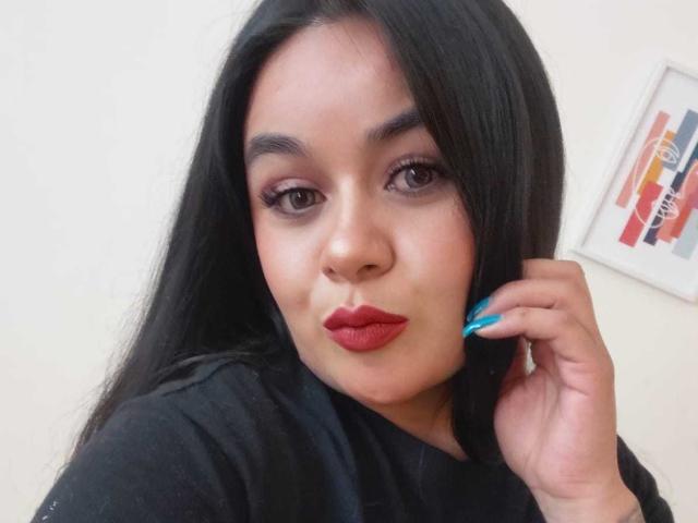 HannahMolly with long black hair, red lipstick, and blue nails, pouting her full ass while touching her ear, wearing a black shirt, in a selfie.