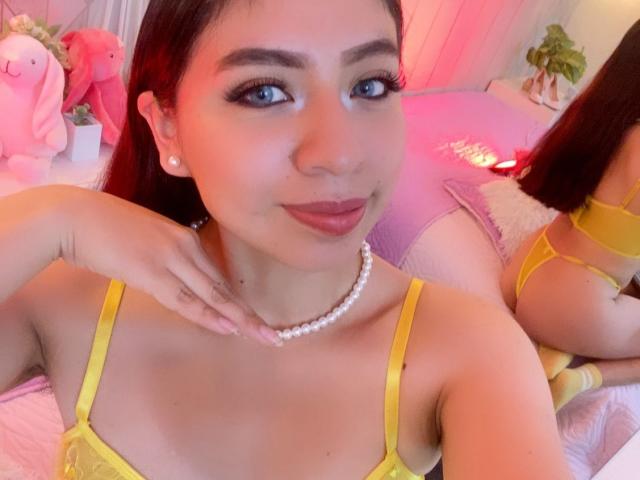 ZoeRedds with yellow strap dress, pearl necklace, and red lipstick, showing off her tits in a mirror selfie, pink bed, and stuffed animals in the background.