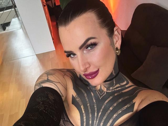 KathaleaNoir-hot, tattooed chest, purple lipstick, nose ring, dark hair, green eyes, black mesh sleeves, smiling, wooden floor, black couch, gold earrings, arm tattoos.