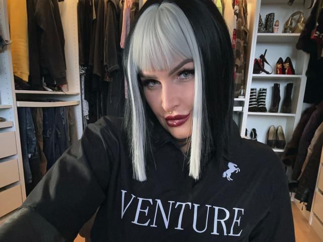 KathaleaNoir-hot with black and white hair, nose ring, and dark lipstick, wearing a black "VENTURE" shirt, standing in a closet with shoes and clothes.
