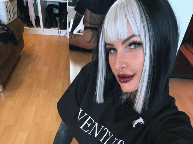 KathaleaNoir-hot with white and black bob, nose ring, dark lipstick, and black Venetia hoodie, sitting on wooden floor, smiling at camera. Boobs covered by hoodie.