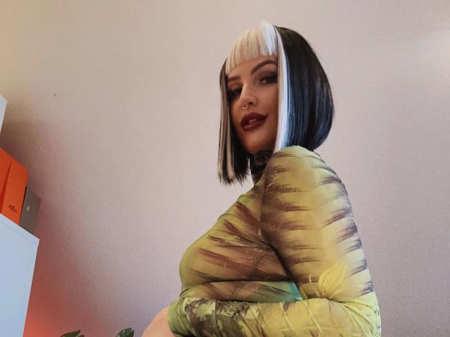 KathaleaNoir-hot with a black and white bob, nose ring, dark lipstick, wearing a green and yellow tie-dye top, showing her tits. Her ass is slightly visible.