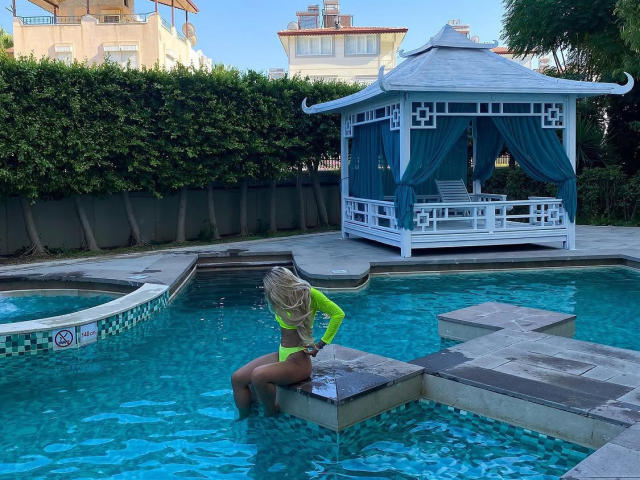 LilianaWax with blonde hair, neon green long-sleeve top, and green thong, sitting on pool ledge, blue water, white gazebo with green curtains, backyard, clear sky.