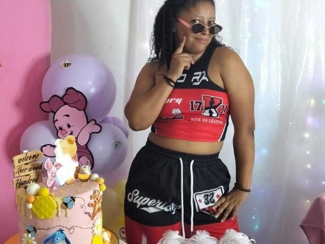  FranciSquirtt stands in front of a decorated table. She's wearing a red and black sports crop top with "BAY" and "3" logos, and matching black and red athletic shorts. She has curly hair, glasses, and a finger to her lips. The table has a pink cake with yellow and blue decorations, a "Happy Birthday" sign, and a Hello Kitty balloon cluster. The background is a white curtain with colorful sparkles. She's slightly to the right, with the table on the left.