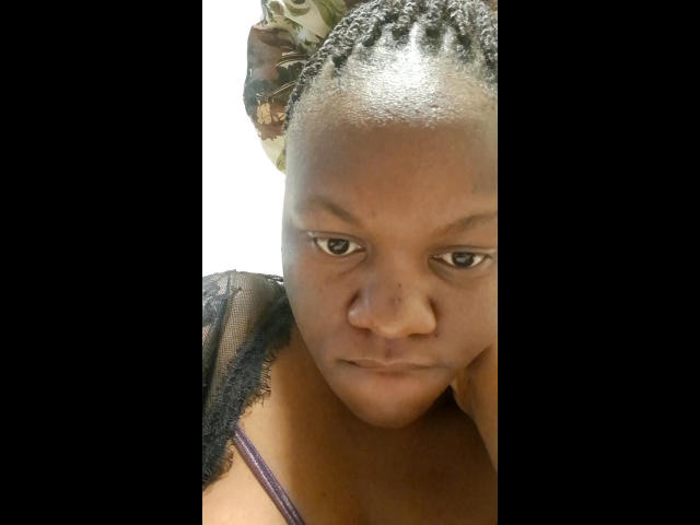 EbonyBeauty69 has a serious expression, small nose, and short braided hair with a floral headwrap. She's wearing a black lace top with purple straps.