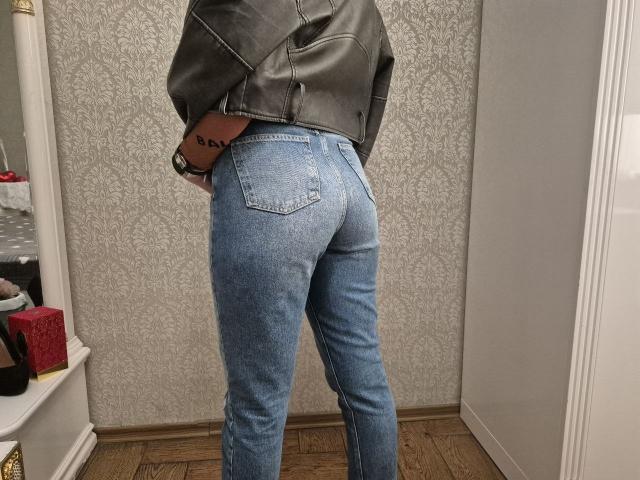 LoriEla's ass in tight blue jeans, wearing a grey leather jacket, standing in a room with wooden floor, tattoo on right wrist, wristwatch on left wrist.