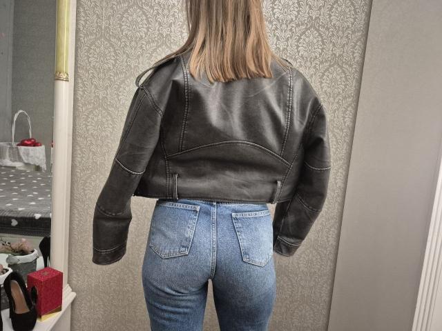 LoriEla's back, wearing a black leather jacket and blue jeans, showing her tight ass. She has straight, blonde hair and stands in a room with patterned wallpaper.