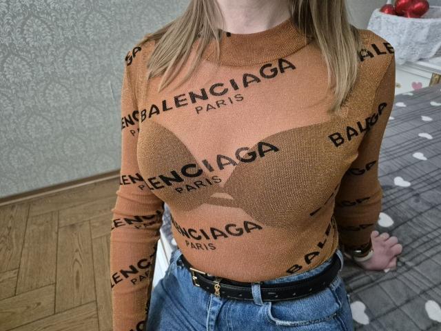 LoriEla wearing a brown, long-sleeve, patterned top with "Balenciga Paris" text, highlighting her big tits, and blue jeans with a black belt. Blonde hair, wooden floor.