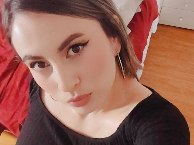 AmiiRosse with pale skin, brown eyes, and brown hair, wearing black and looking at camera, showing small tits, wearing thin gold hoop earrings, in a room with red bed.