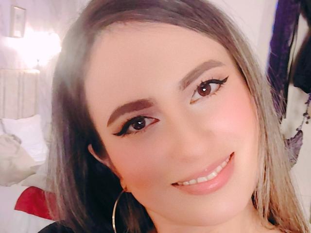 AmiiRosse has brown eyes, long brown hair, and a pink smile with winged eyeliner. She's smiling at the camera with her tits partially visible. She's wearing large hoop earrings.