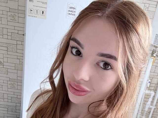KarinaVibe69 has puffy, pinkish-lipsticked lips and brown eyes with long, wavy, light brown hair, looking at the camera with a slight, cute smile.