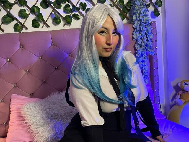 MaddieLesYoux has long, white-to-blue gradient hair, wearing a white shirt with a black necktie and black suspenders. She's sitting on a pink, tufted headboard bed with a fluffy white pillow and a pink pillow. The headboard has green vines and small lights. A stuffed toy is on the right. Her makeup includes pink eyeshadow and lipstick. The lighting is blue. The image is vibrant and detailed.