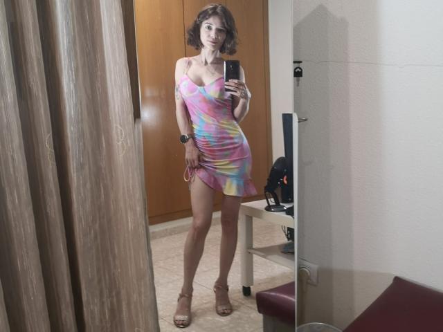 SonyaLovelace with curly brown hair, wearing a colorful, tie-dye, one-shoulder dress, showing her tits, standing in high heels, taking a mirror selfie in a room.