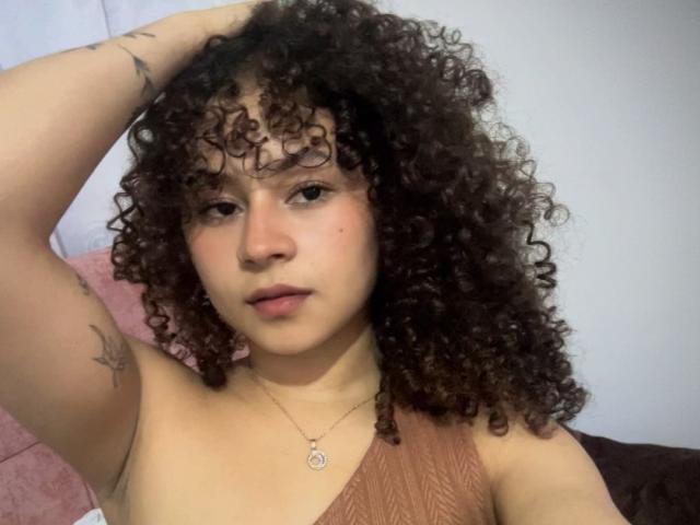 SofiaPassionne with curly brown hair, fair skin, small tattoos on arms, wearing a one-shoulder brown top, hand in hair, neutral expression, wearing a necklace with a circular pendant.