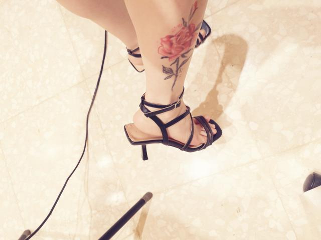 SonyaLovelace's tattooed leg with a red rose, wearing black strappy high heels, standing on a marble floor with a black cord and metal pole in the bottom left.