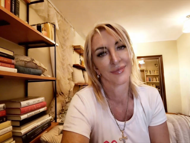 RoseQMagic is a blonde woman with straight, shoulder-length hair and light makeup. She's wearing a white t-shirt with a graphic and a silver necklace with a cross pendant. She has a slight smile and is looking at the camera. Her skin is fair, and she's wearing small earrings. The background shows a bookshelf with stacked books and a curtain. The room is warmly lit with beige walls. The image has a casual, candid feel.