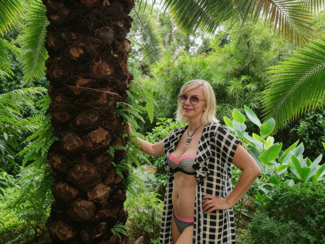 RoseQMagic stands next to a palm tree, wearing a black and white checkered open robe over a green and pink striped bikini. She has blonde hair, sunglasses, and a necklace. Her right hand rests on the tree. The background is dense with green foliage. The palm tree has rough, textured bark. The overall composition is natural and relaxed, with a focus on RoseQMagic's casual, confident stance.