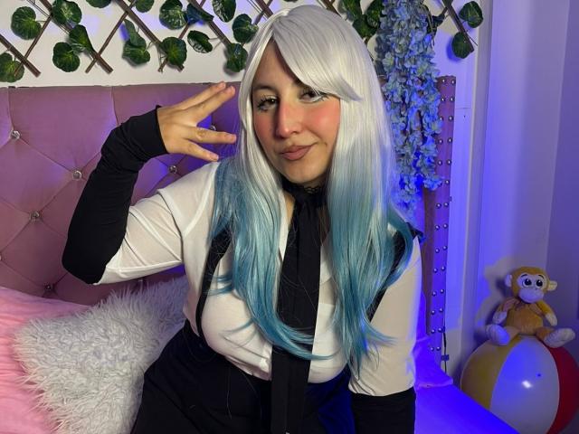 MaddieLesYoux with white and blue hair, smiling, wearing a black necktie, white shirt, and black sleeves, making a peace sign, pink tufted headboard, fluffy white pillow.
