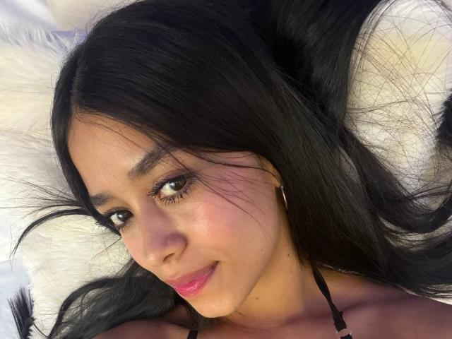 Close-up photo of LaurentRay lying on a white, fluffy surface. LaurentRay has long, straight black hair, brown eyes, and a slight smile. Her skin is smooth with a light brown tone. She wears small gold hoop earrings and has pink lipstick. Her eyelashes are long and dark. The light highlights her cheekbones and forehead. The image focuses on her face, showing her relaxed and comfortable expression. The soft texture of the background contrasts with her sleek hair.