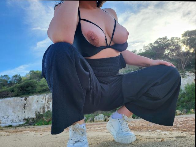 Linnea-hot crouching, wearing a black strappy top exposing her big, round boobs with visible nipples, black pants, and white sneakers, outdoors with a blue sky and trees.