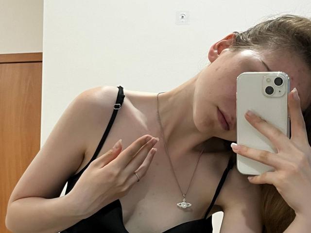 DeeannMilloy taking a selfie, wearing a black bra, holding an iPhone, with a silver necklace, light skin, blonde hair, and a ring on her right hand.