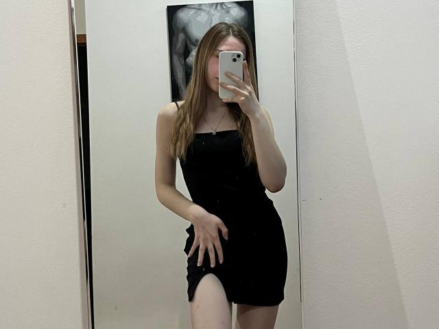 DeeannMilloy with long brown hair, wearing a tight black dress, showing a thigh cutout, holding a phone, covering her face, standing in a mirror selfie.
