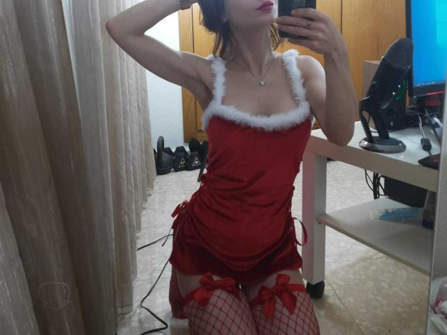 SonyaLovelace in red Santa dress with white fur trim, red fishnet stockings, and red garters, kneeling, taking mirror selfie, showing her tits and ass.