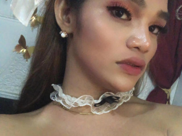 Close-up of EsaMarie's face with a slight smile. She has light brown skin, full lips with pink lipstick, and subtle eye makeup with pink eyeshadow. Her brown hair is partially visible. She wears a white lace choker with a small black bow, and pearl earrings. Her eyebrows are neatly shaped. The texture of her skin is smooth, with a slight sheen. The background includes a white wall with butterfly decorations and a red curtain. The image is a selfie.