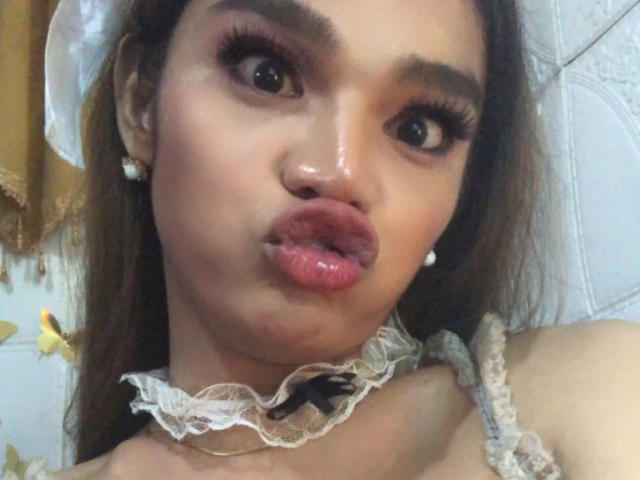 Close-up of EsaMarie with light brown skin and long brown hair, pouting her pink glossy lips. She wears a lace choker with a small black bow and pearl earrings. Her dark brown eyes are wide, and she has thick, long eyelashes. The texture of her skin is smooth, and her makeup is subtle. The image is focused on her face, which takes up most of the frame.