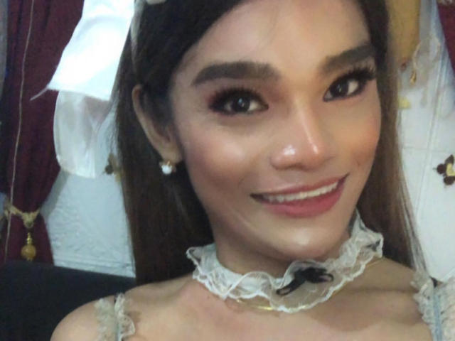 Close-up of EsaMarie, a young woman with light brown skin and long brown hair. She has dark eyebrows, long eyelashes, and is smiling with red lipstick. She wears a white lace choker with a small black bow, and pearl earrings. Her makeup is subtle with pink eyeshadow. The background includes red curtains with gold tassels. The image has a soft, slightly blurry quality.