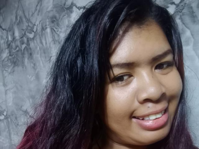 Close-up of MariahSquirting69 with dark, wavy hair that transitions to maroon at the ends. She has a light brown skin tone, small nose, and a slight smile showing her teeth. Her eyebrows are thick and natural. The texture of her hair is slightly messy. She has a small nose piercing on the right side. The background is a gray marble pattern. The image has a soft, natural light. MariahSquirting69's eyes are looking slightly to the right.